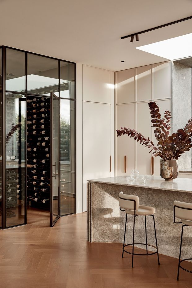 A refined contemporary kitchen and entertaining space with a large stone island and integrated seating. Full-height pale cabinetry with slim timber-toned handles lines the rear wall, while a black-framed glazed wine room is visible to the left. Warm herringbone timber flooring, dark metal-framed glazing, a skylight, and soft natural light create an elegant, luxurious atmosphere.