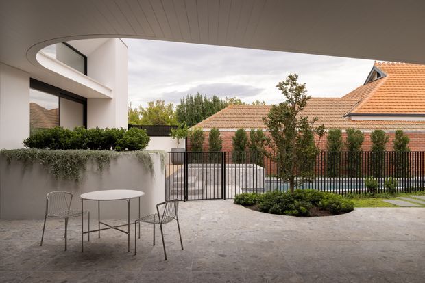 A contemporary covered outdoor courtyard or terrace viewed from beneath a curved, lined soffit. The space has a pale concrete or stone-paved surface, minimalist outdoor dining furniture, raised rendered planter beds with clipped shrubs and trailing greenery, and a black metal picket fence. A modern white multi-level building is visible on the left, while an adjacent traditional brick house with terracotta tiled roofs forms the backdrop.
