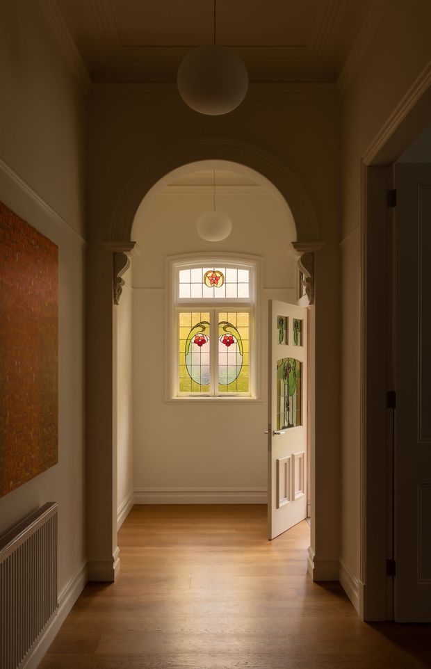 A warmly lit traditional residential entry hall viewed from inside, with polished timber flooring, cream painted walls, decorative cornice and skirting, a high coffered ceiling, and a large arched opening framed by ornate trim. At the far end is a partly open front door with distinctive stained-glass panels and a matching stained-glass window. A classic radiator and framed artwork line the hallway, while spherical pendant lights hang centrally.