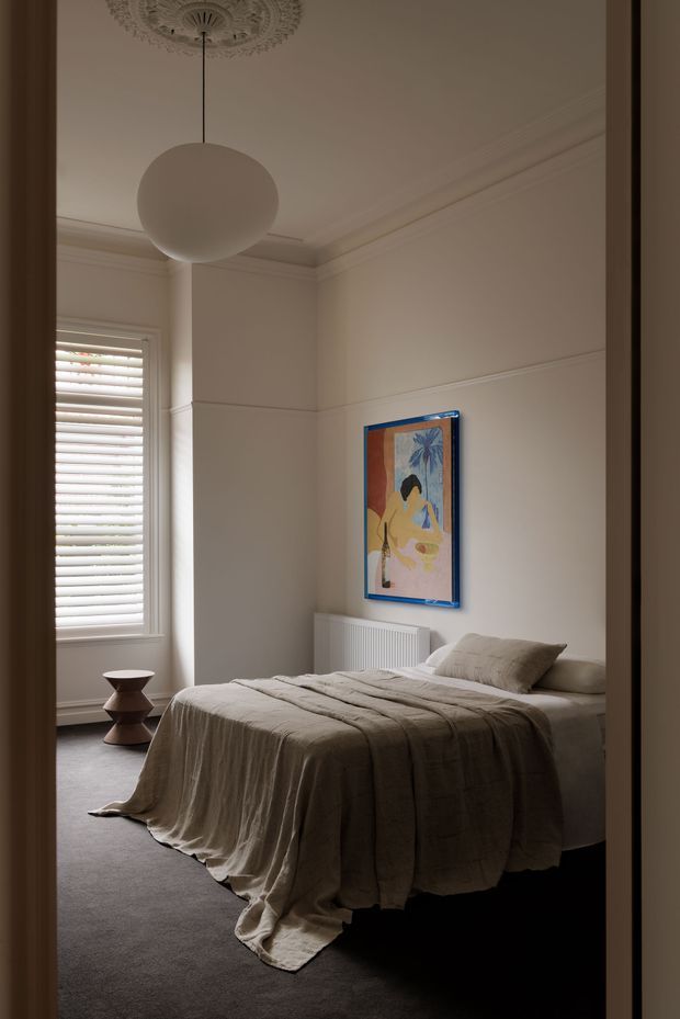 A softly lit bedroom viewed through a doorway, featuring a neatly made bed with a taupe linen coverlet, a sculptural bedside table, framed artwork, a white radiator, plantation shutters, ornate ceiling detailing, decorative cornices, and a large spherical pendant light. The room has a calm, refined traditional shell with restrained contemporary furnishings.