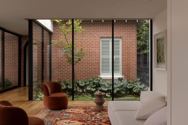 A warm, refined living room viewed through expansive black-framed glazing overlooking an internal brick courtyard. The room features a light timber floor, a patterned oriental-style rug, a white upholstered sofa, sculptural rust-coloured lounge chairs, and a small decorative side table. The courtyard has red brick walls, lush planting, and a shuttered window, creating a strong indoor-outdoor connection.