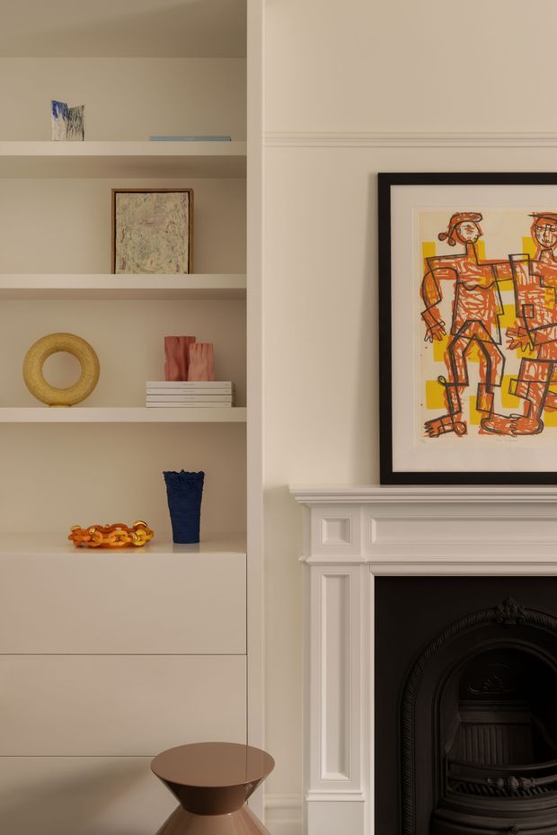 A close-up view of a refined living room wall featuring bespoke built-in shelving and a traditional white painted fireplace mantel. The shelves display curated artwork, ceramics, books, and decorative objects, while a framed figurative artwork rests above the fireplace. The room uses warm neutral tones with classic architectural detailing and a dark firebox.