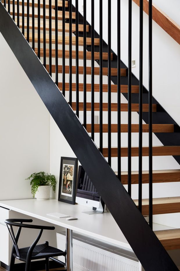 Interior view of a contemporary open-riser staircase with warm timber treads, black metal stringers and vertical balustrade rods. A compact white desk and computer workspace sits beneath the stair, with a black chair, framed artwork and potted plant. The space has white walls and a clean, light-filled minimalist character.