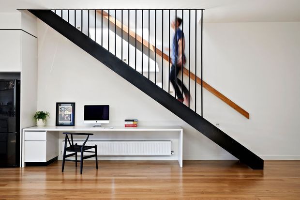 Bright contemporary interior featuring a straight staircase with a bold black steel stringer and slender vertical balustrade, timber handrail, white walls, and warm timber flooring. A slim white built-in desk runs beneath the stairs with a computer, framed artwork, chair, books, and radiator. The space appears open, minimal, and multifunctional, with a person moving along the stairs.