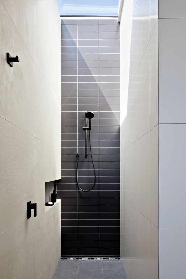A narrow, contemporary walk-in shower with a tall skylit opening. Large pale wall tiles line both side walls, while the rear wall is finished in dark grey horizontal subway tiles with white grout. Matte black shower fittings, controls, and a recessed niche provide contrast. A linear floor drain and grey tiled floor complete the minimalist bathroom design.