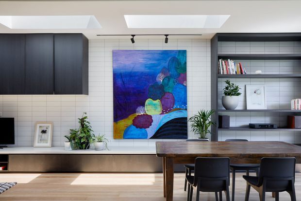 Bright contemporary dining and living area with a long dark timber dining table and black chairs in the foreground. The rear wall combines white horizontal tile cladding, dark built-in cabinetry, open shelving, a low media console, plants and a large colourful abstract artwork. Two skylights and track lighting provide strong natural and artificial illumination.