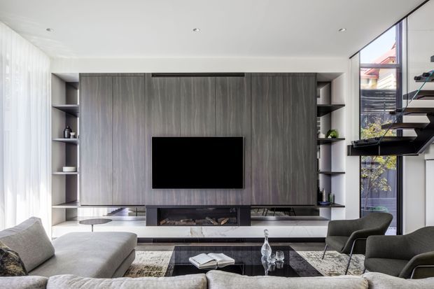Contemporary living room with a large grey timber-look built-in media wall, open shelving, a central television and linear fireplace. A low coffee table, sectional sofa and upholstered lounge chairs sit on layered rugs, while a glazed opening and floating staircase bring in natural light.