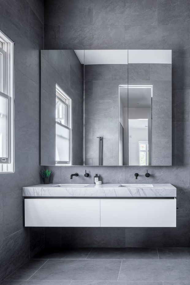 A contemporary double vanity bathroom with dark grey stone-look wall and floor tiles, a white marble-look benchtop, white floating cabinetry, black tapware, and large mirrored cabinetry above the vanity. Natural light enters through a narrow window, creating a cool, refined monochromatic interior.
