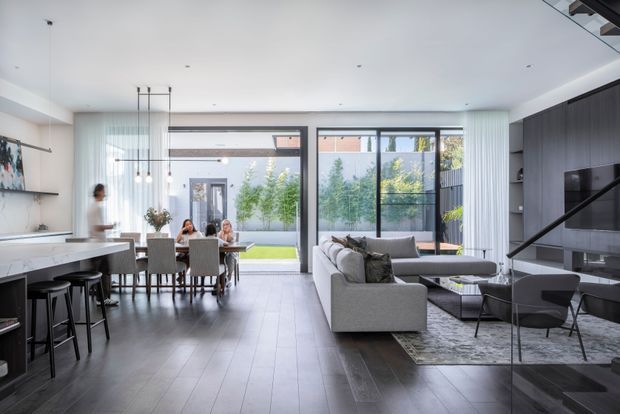A bright, open-plan contemporary interior combining a kitchen, dining area and living room. The space features dark timber-look flooring, a large island with bar seating, a dining table, a grey sectional sofa, extensive floor-to-ceiling glazing, and a dark built-in media wall with diagonal detailing. The restrained monochromatic palette is softened by garden views and natural light.