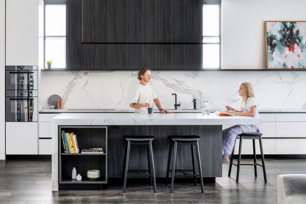 Bright contemporary kitchen with a large central island, dark vertically grained cabinetry, white stone surfaces, a veined stone splashback, integrated appliances, and black bar stools. Two people are seated and standing at the island, with abstract artwork and understated styling adding visual interest.