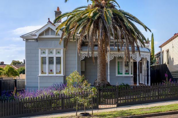 Street-facing view of a single-storey heritage-style cottage with a pale grey weatherboard facade, decorative gabled frontage, ornate verandah posts and fretwork, timber-framed windows with shutters, and a prominent palm tree in the front garden. A dark picket fence and landscaped planting define the property boundary.