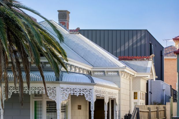 Exterior view of a traditional heritage-style house with ornate white lacework verandah detailing, a corrugated metal pitched roof, decorative gable trim, chimney, and pale cladding. A modern dark metal-clad extension is visible behind the original dwelling, with a palm tree in the foreground.