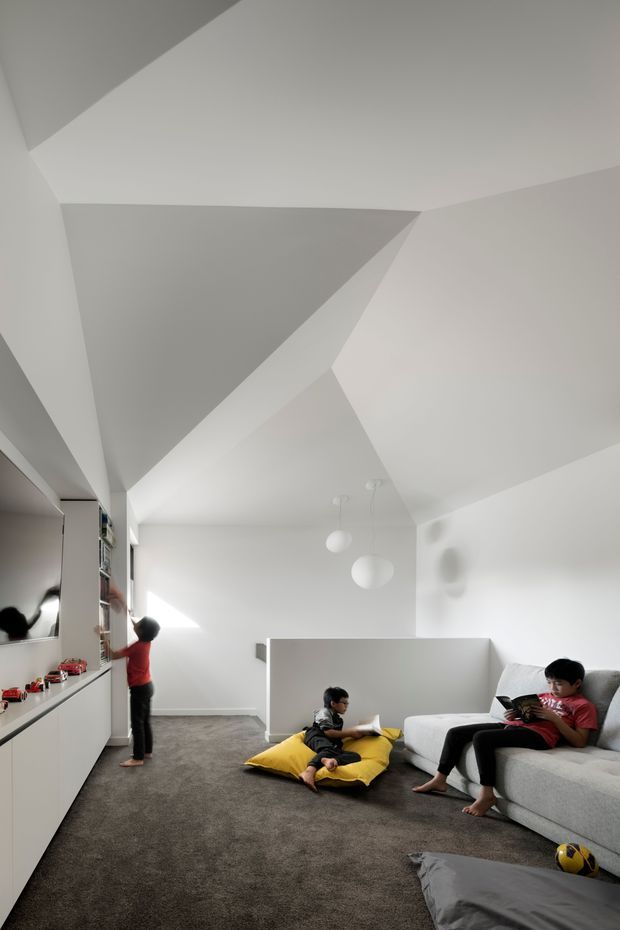 Bright, minimalist family living area with a dramatically faceted angular white ceiling, white walls, grey carpet, built-in white cabinetry and entertainment storage along the left wall, and a large light-grey sectional sofa. Two children are reading and playing, with additional floor cushions and a ball creating a relaxed family atmosphere. Sculptural white pendant lights hang near the centre of the room.