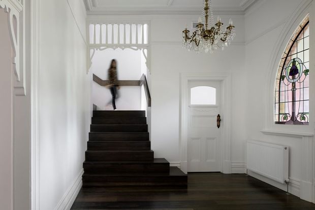 A bright, refined traditional entry hall with a dark timber staircase, white panelled walls and ceiling, ornate chandelier, decorative leadlight window, and a white front door. A blurred person is moving on the stairs.