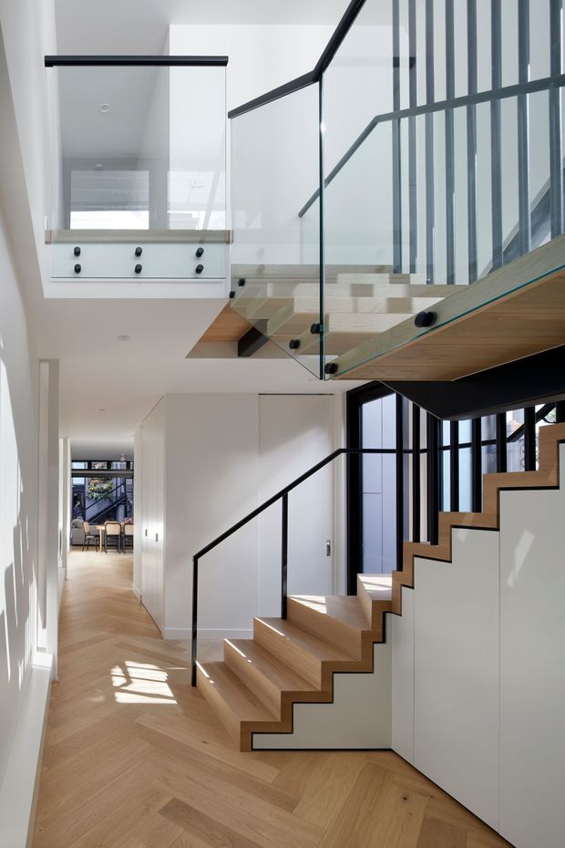 A bright, contemporary interior focused on a sculptural staircase and upper-level landing. The staircase has light timber treads and matching herringbone timber flooring, white zigzag stringer walls, clear glass balustrades, and slim black metal handrails. Full-height black-framed glazing lines the stairwell, while a hallway extends toward a dining area in the background. The space is airy and vertically connected, with clean white walls and strong natural light.
