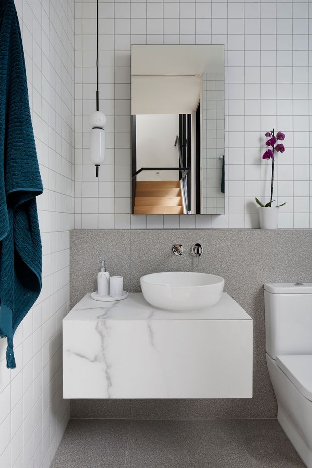 A compact contemporary bathroom with white square wall tiles, a grey speckled tiled vanity wall and floor, and a floating white marble-look vanity with a round vessel basin. A large rectangular mirrored cabinet reflects a stairway, while a white toilet, wall-mounted tapware, teal towel, pendant light and potted orchid add detail.