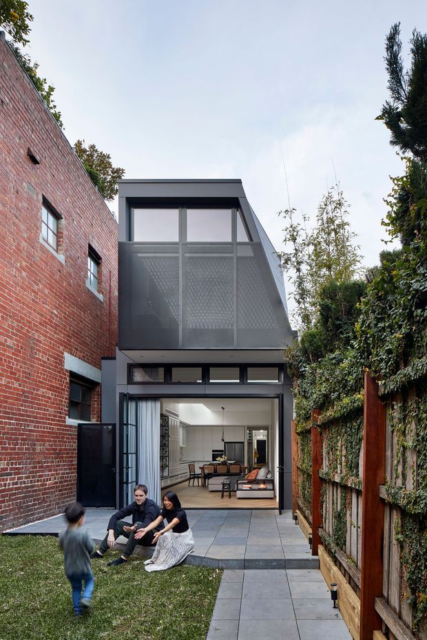 A narrow urban courtyard showing a contemporary multi-level rear extension between an exposed red-brick wall and a timber garden fence. The extension has dark-framed glazing, a projecting upper volume with perforated metal screening, and an open ground-floor living area connected to a paved terrace and lawn.