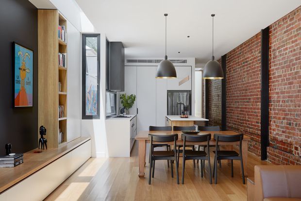 Open-plan contemporary dining and living area with a timber dining table and dark chairs beneath two oversized pendant lights. The room features light timber flooring, white kitchen cabinetry, a dark built-in media wall with shelving, exposed red brick walls with black steel framing, and tall narrow windows. A compact kitchen is visible at the rear, while a leather sofa edge appears in the foreground.