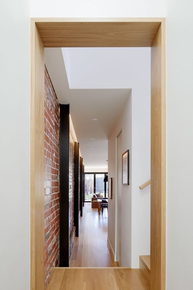 A bright, narrow residential hallway viewed through a substantial light timber-framed opening. The corridor features exposed red brickwork along the left wall, white plaster walls and ceiling, light timber flooring, recessed downlights, framed artwork, and a timber handrail beside a short stair. A living area with seating and full-height glazing is visible at the far end. A rooflight or skylight brings additional daylight into the passage.