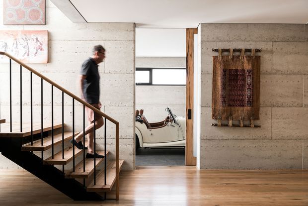 Interior view of a contemporary home featuring an open timber-and-black-metal staircase, polished concrete walls, warm timber flooring, and a wood-framed opening into a room with a vintage car. A woven textile wall hanging and artwork add decorative detail.