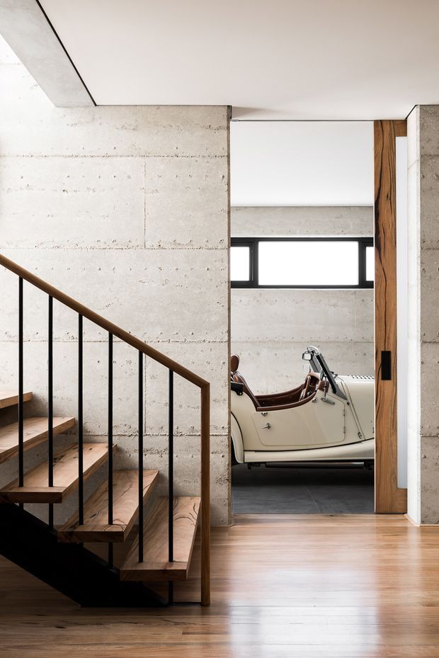 Interior view of a contemporary concrete and timber home, focused on a staircase and hallway opening toward a garage or display area. The staircase has warm timber treads and handrail with slim black metal balusters, while exposed board-formed concrete walls and ceiling create an industrial architectural character. A vintage cream-colored car is visible through the opening, with a narrow horizontal window beyond.