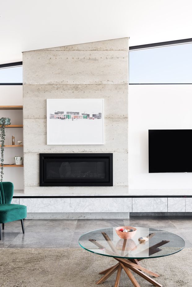Bright contemporary living room featuring a full-height pale concrete fireplace surround with a black built-in firebox, a wall-mounted television, narrow clerestory glazing, open timber shelving, and a long low marble-faced media cabinet. A teal upholstered armchair, round glass coffee table with timber starburst base, and neutral rug complete the refined interior.
