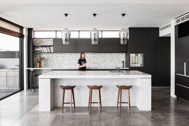 Contemporary kitchen dominated by a large light stone island with seating for three, dark flat-panel cabinetry, a white brick-tile splashback, and three pendant lights. Full-height glazing and a dark polished floor contribute to an open, refined interior.