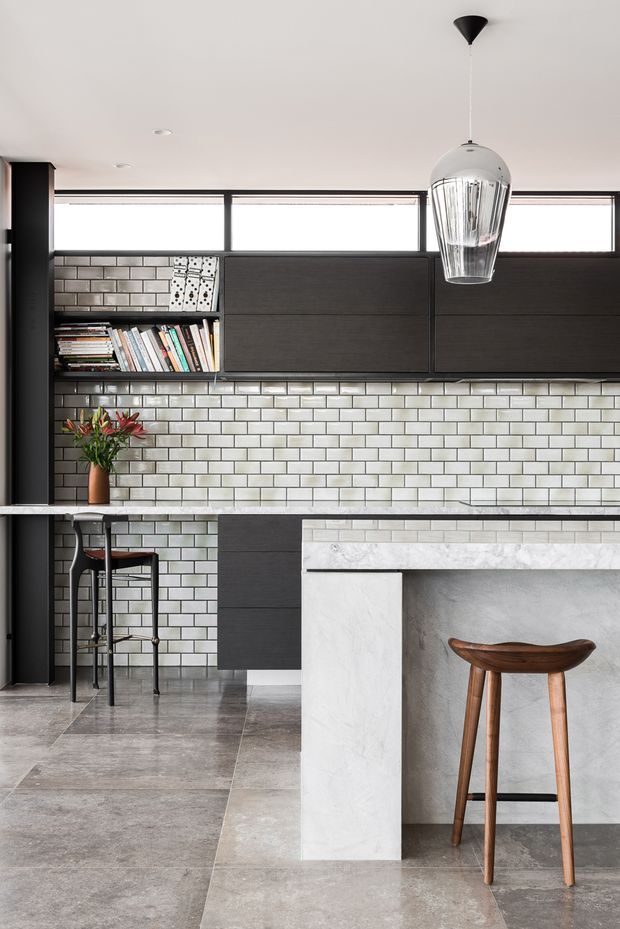 A contemporary kitchen with a compact breakfast peninsula and a larger foreground island. The space features dark charcoal flat-panel cabinetry, a full-height glossy light subway-tile splashback, pale stone-look benchtops, grey large-format floor tiles, slim clerestory-style windows, and a sculptural clear glass pendant light. Open shelving holds books and a vase of flowers, while timber bar stools add warmth.