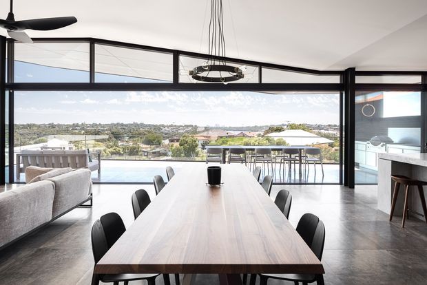 A contemporary open-plan dining and living space with a long timber dining table, black dining chairs, polished concrete flooring, and expansive black-framed glazing. The glazing opens toward an outdoor terrace with a pool and a broad suburban landscape. A kitchen or bar area is partially visible at right, while a large sectional sofa and ceiling fan sit to the left. A sculptural pendant light hangs above the table.