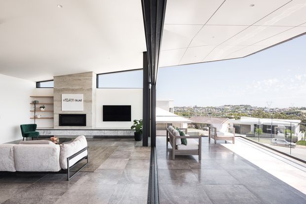 Bright contemporary open-plan living room with a substantial stone-clad fireplace wall, wall-mounted television, built-in shelving, low-profile upholstered seating, and a large glazed opening leading to an outdoor terrace. Full-height dark-framed glazing reflects the living area and provides views toward a suburban hillside. The room has a polished stone or large-format tiled floor, white ceiling, and a restrained neutral palette.
