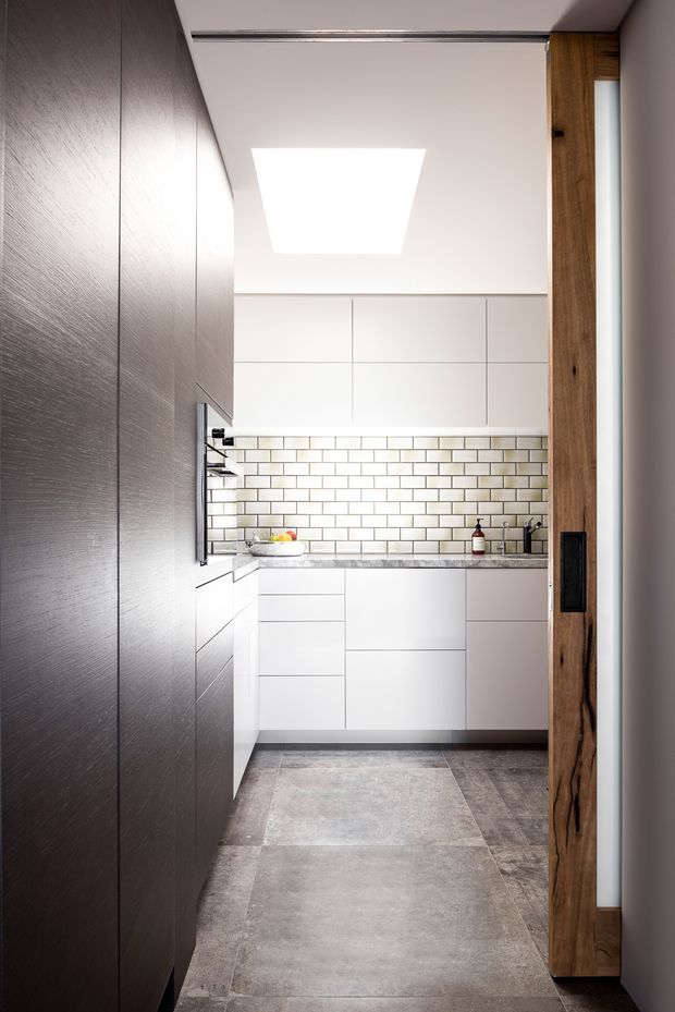 Compact contemporary kitchen viewed from the entry, with floor-to-ceiling dark textured cabinetry on the left, a white handleless kitchen run at the back, white subway-tile splashback, pale stone-look benchtop, and large grey floor tiles. A warm timber-framed doorway contrasts with the restrained white, grey and dark palette. A large rectangular ceiling light/skylight brightens the space.
