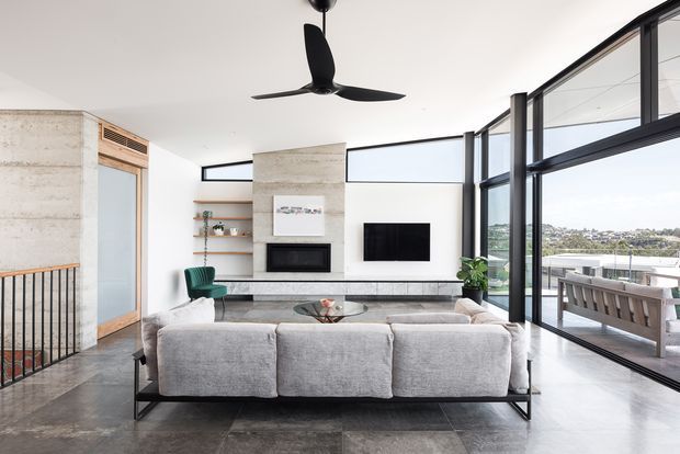A bright, contemporary open-plan living room with a large light-grey sectional sofa facing a minimalist white fireplace and wall-mounted television. The room features polished dark grey floor tiles, exposed concrete feature walls, timber detailing around a doorway and shelving, expansive black-framed floor-to-ceiling glazing, a black ceiling fan, and a balcony or terrace with a view beyond.