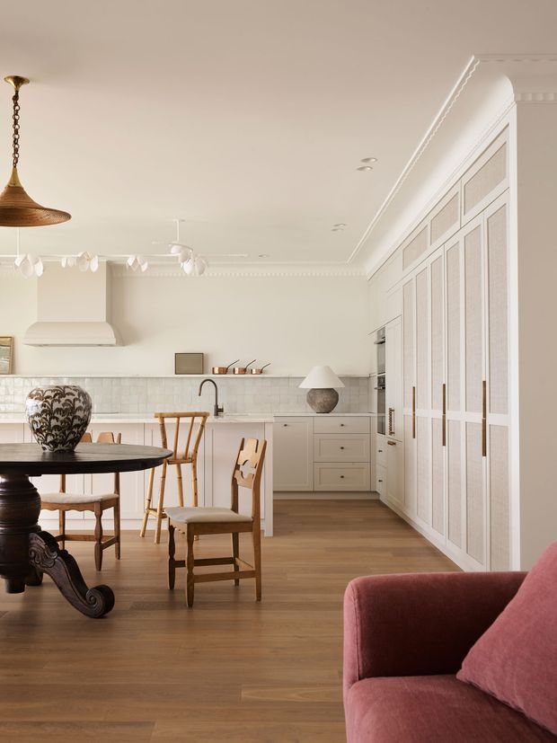 A refined open-plan kitchen and dining area with warm timber flooring, soft off-white walls and ceiling, detailed traditional mouldings, bespoke pale cabinetry, and a large built-in wall of textured-panel cupboards. The kitchen includes a tiled splashback, island bench, integrated rangehood, and brass-toned fixtures. A dark carved timber dining table, woven pendant light, upholstered dining chairs, and a muted rose armchair add layered, elegant character.