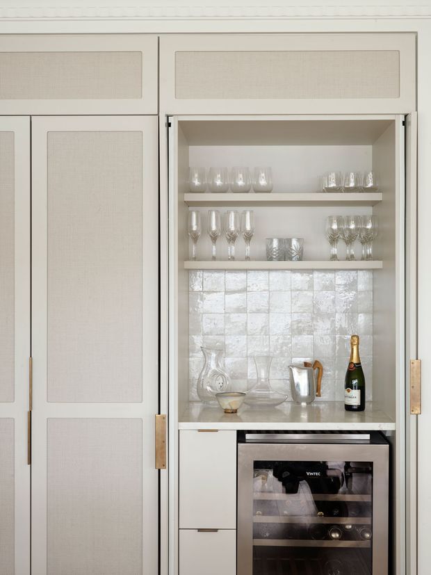 Built-in home bar or drinks cabinet with pale painted cabinetry, inset textured fabric-like door panels, brass-toned hardware, open glassware shelving, reflective silver-white tiled splashback, light stone benchtop, and an integrated wine fridge below. The styling is refined and light, with champagne and decanters displayed.