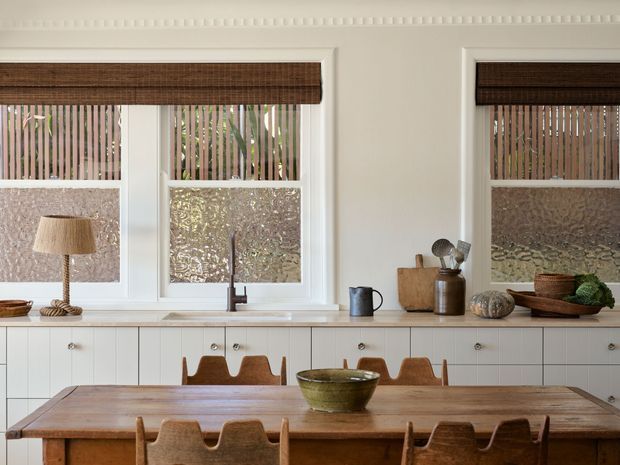 Warm, carefully styled kitchen and dining area with white shaker-style cabinetry, a timber dining table and chairs, three large windows with woven Roman blinds, and a simple farmhouse-inspired decorative arrangement on the countertop. The room has abundant natural light, timber accents, and a calm, refined atmosphere.