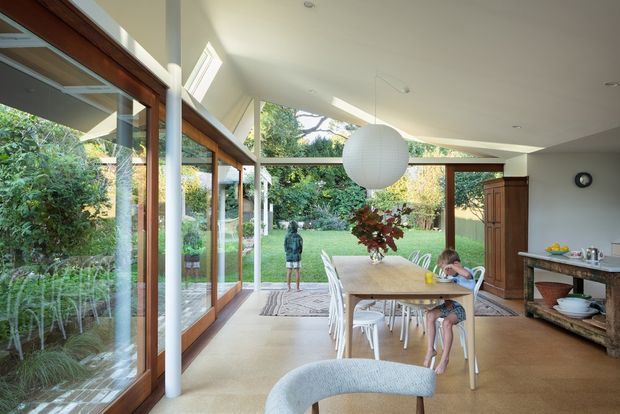 Bright open-plan dining area with extensive timber-framed glazing opening onto a lush garden. A long timber dining table with white chairs sits beneath a large spherical pendant light, while a dark timber sideboard and stone-topped console occupy the right side. The space has a pale neutral palette, polished concrete or stone-like flooring, white walls, and a strong indoor-outdoor connection.