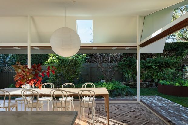 A bright, glazed dining area opening onto a landscaped garden. The space features a long timber dining table with curved-back chairs, a large spherical paper pendant light, extensive windows and doors, white structural framing, timber detailing, and a patterned area rug over a dark floor.