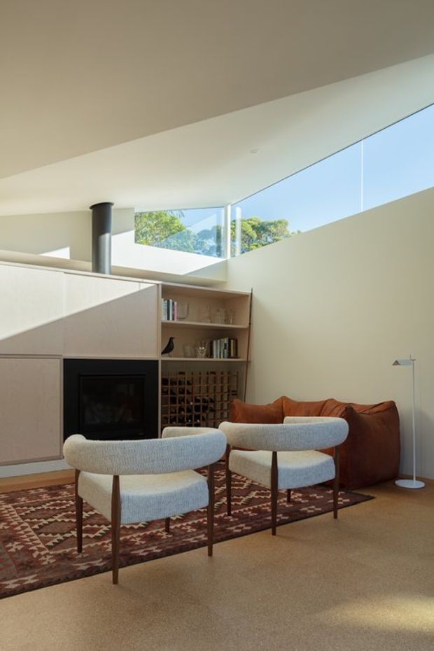 A warm, contemporary living room with a sculptural built-in fireplace, integrated timber shelving and wine storage, two upholstered lounge chairs, a brown sofa, and a patterned area rug. High clerestory glazing and skylight-like windows bring in abundant natural light, while a dark cylindrical flue and low-profile floor lamp add architectural and decorative interest.