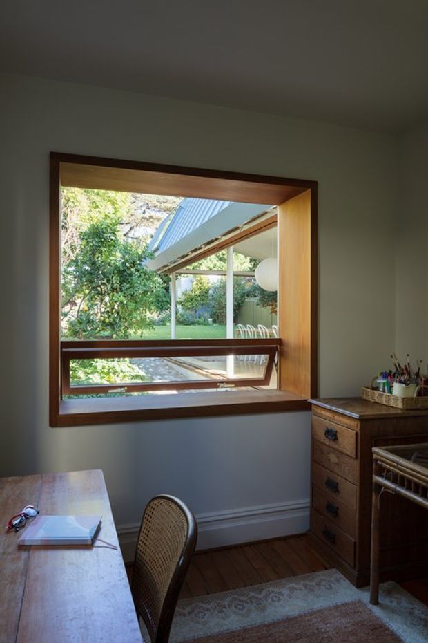 A compact, warmly lit home office or study with a large timber-framed awning window overlooking a leafy garden and covered outdoor area. The room contains a wooden writing desk, woven-back chair, vintage wooden chest of drawers, stationery supplies, a small rug, and dark timber flooring. The window provides the main architectural feature and introduces abundant garden light.
