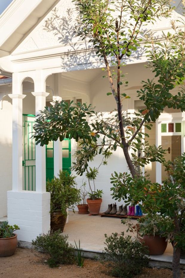 A bright, sheltered front entry porch or courtyard of a traditional rendered home, framed by white classical columns and arched openings. The facade features decorative mouldings, a covered roof structure, and a glazed entrance door with vivid green geometric framing and coloured panes. Potted plants and established landscaping surround the tiled or concrete entry, with shoes arranged near the doorway.