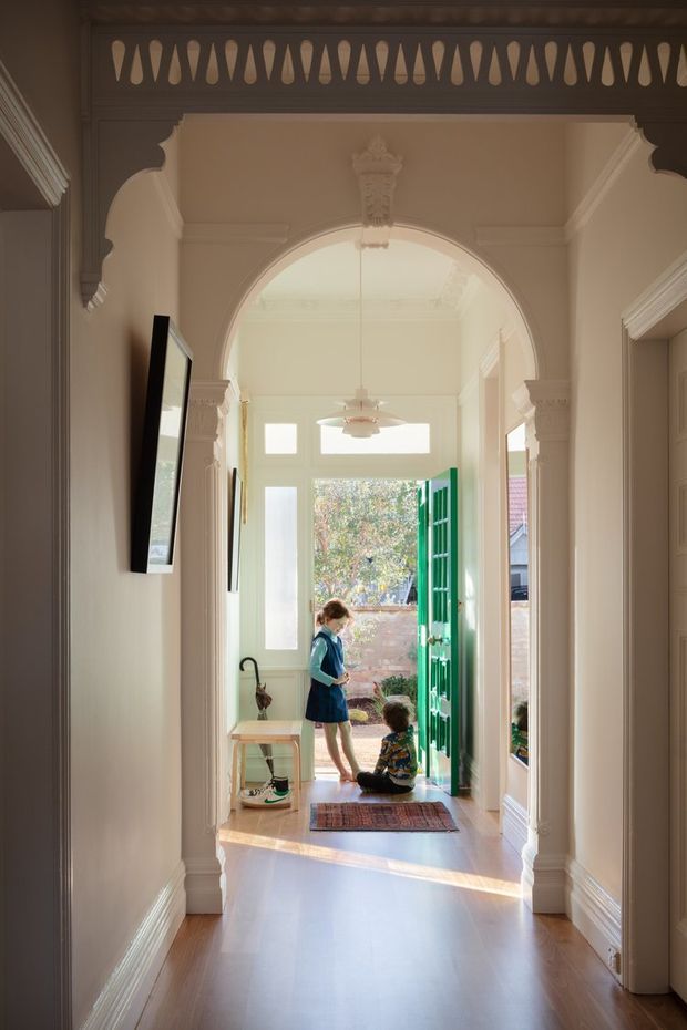 A bright, ornate residential hallway and entryway viewed from inside, featuring a polished timber floor, detailed classical archway, decorative plaster mouldings, columns, and a perforated timber screen above the opening. The hallway leads to a sunlit front entrance with a vivid green door and glazed sidelights, with children near the doorway. A large wall mirror reflects the entry area.