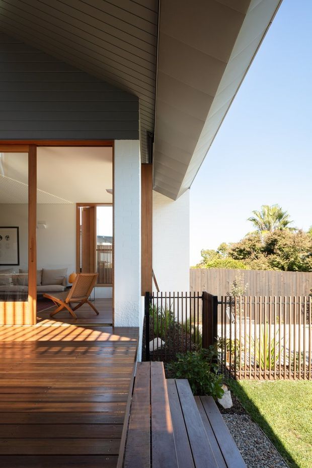 Exterior view of a contemporary residence showing a covered timber deck and adjoining outdoor bench, with white masonry walls, timber-framed sliding glazing, dark horizontal cladding beneath the roof, and a fenced landscaped yard. The open glazing reveals part of a living room inside. Strong roof overhangs and warm timber surfaces define the sheltered outdoor space.