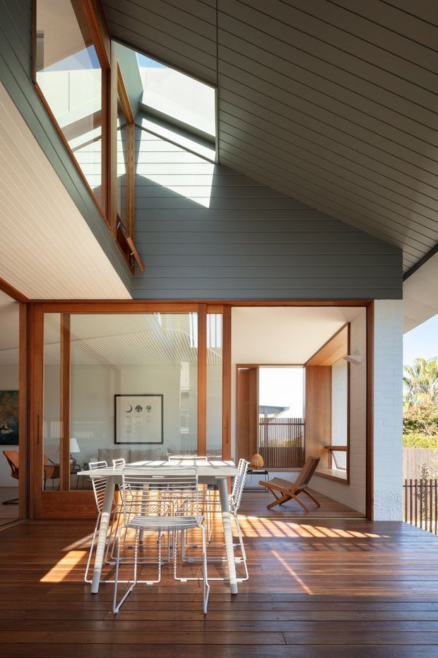 Sunlit modern indoor-outdoor dining area opening onto a timber deck. The space features extensive timber-framed glazing, a high sloping ceiling with dark horizontal lining, clerestory/skylight-like glazing, timber wall and ceiling accents, and a white dining table with wire-frame chairs. A separate lounge chair is visible inside, with strong sunlight creating geometric shadows across the timber floor and deck.