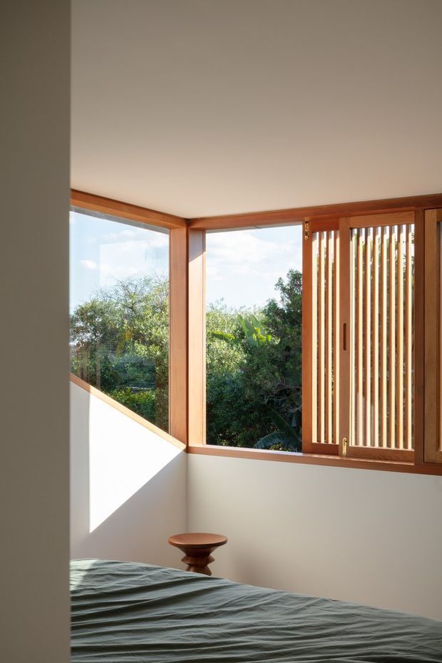 A serene bedroom with a low-profile bed in the foreground, warm timber-framed corner windows, and a vertical timber louvred sliding screen. Natural greenery is visible outside, while sunlight creates a strong triangular patch of light on the pale wall. The room has a simple, restrained design with warm wood detailing and muted bedding.