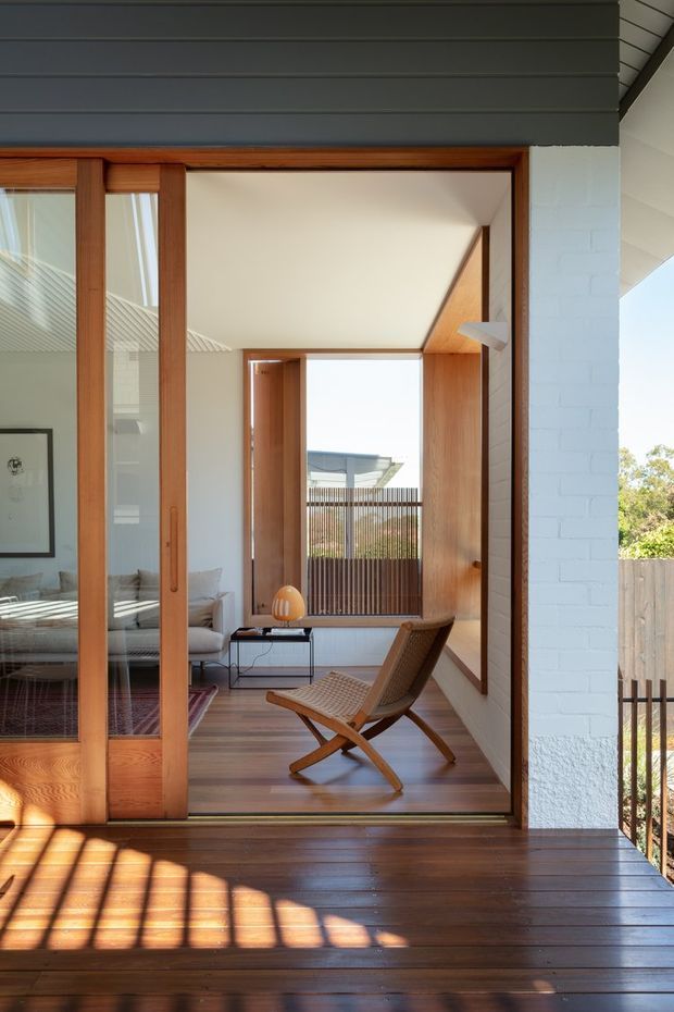 A bright architectural view from a timber deck into a refined indoor-outdoor living area. The composition features large timber-framed sliding glass doors, white-painted brickwork, warm timber flooring and wall lining, a woven lounge chair, and a glimpse of a living room with a sofa and framed artwork. Strong sunlight creates linear shadows across the dark timber deck, while a screened outdoor area is visible beyond the interior.