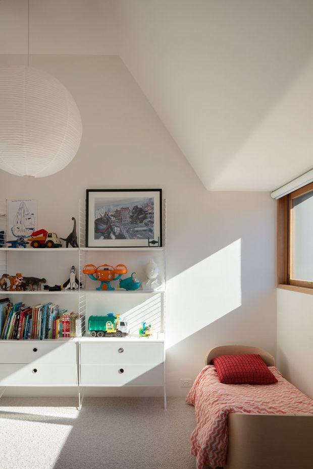 A bright children's bedroom with a single bed beside a large white open shelving and drawer unit filled with books and toys. The room has pale walls, a light carpeted floor, a timber-framed window with a roller blind, a large white paper pendant light, framed artwork, and strong geometric sunlight across the wall and floor. The styling is playful and colourful within a simple, clean architectural shell.