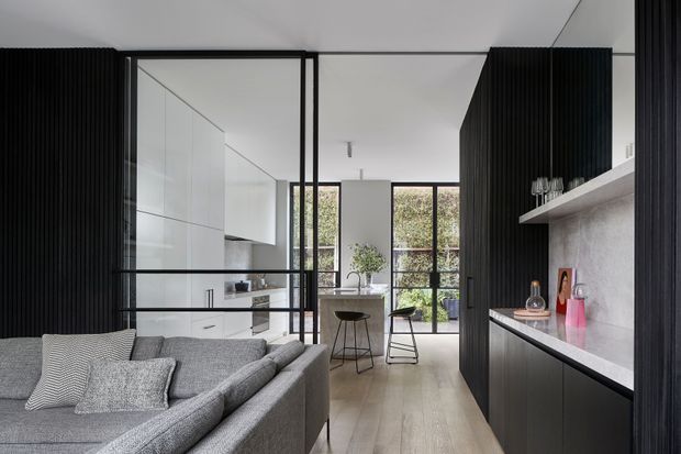 A refined open-plan contemporary interior combining a living room and compact kitchen. A grey sectional sofa occupies the foreground, while black-framed glass partitions separate the living area from a white kitchen and dining zone. The kitchen features dark vertically fluted cabinetry, a pale stone-look benchtop and splashback, integrated white cabinetry, timber-toned flooring, and full-height windows opening toward greenery.