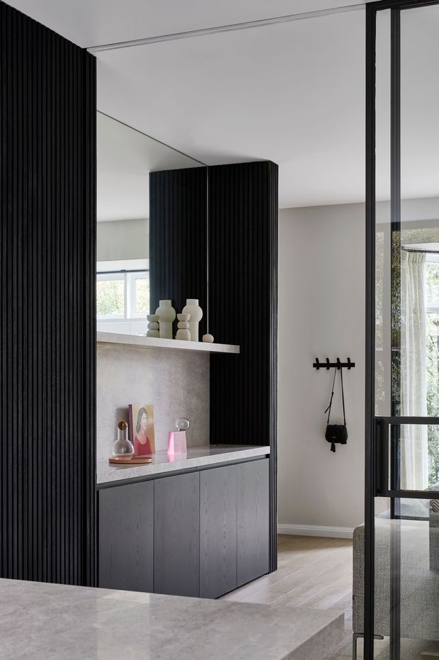 Contemporary interior view featuring a dark vertical fluted timber screen and matching built-in display cabinetry, with a pale stone benchtop and open shelving. Neutral light-grey walls, pale flooring, black-framed glazing, and sculptural decorative objects create a refined, minimalist composition. A hallway or entry area with coat hooks and a glimpse of an adjacent workspace is visible beyond.