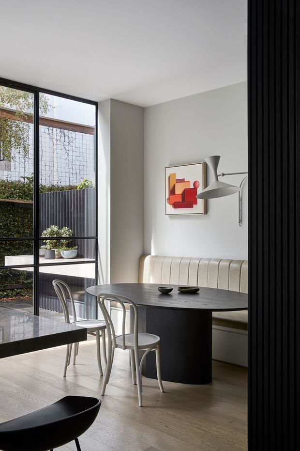 A refined contemporary dining area with a dark oval dining table, curved light-coloured upholstered banquette, and sculptural grey pendant/wall-mounted light. Slim bentwood-style dining chairs surround the table. A large black-framed glazed opening connects the room visually to a planted courtyard, while a black vertical slatted screen frames the right side. Pale timber flooring, soft grey walls, and a colourful abstract artwork create a restrained yet warm interior.