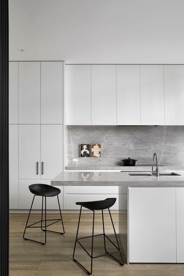 A refined contemporary kitchen with full-height white flat-panel cabinetry, a large grey stone island and matching splashback, integrated appliances, and a slim stainless-steel sink tap. Two black sculptural bar stools sit at the island. The room has light timber flooring, minimal detailing, recessed lighting, and a restrained monochrome palette.